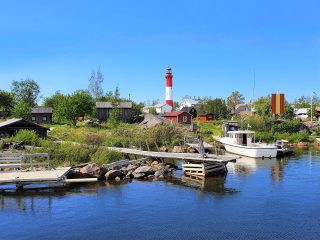 Tankarin majakkasaari kauniina kesäpäivänä. Punavalkea majakka hallitsee kokonaisuutta punaisten ja valkoisten mökkien lomasta. Rannassa on vene.