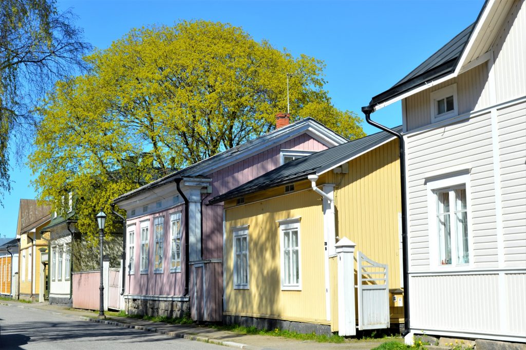 Kokkolan Vanhakaupunki Neristanin pastellinsävyiset talot seisovat kadulla vieretysten. Neristanin kesä on puhkeamassa kukkaan, puiden lehdet ovat juuri vihertyneet ja aurinko luo varjoja vanhoihin rakennuksiin.